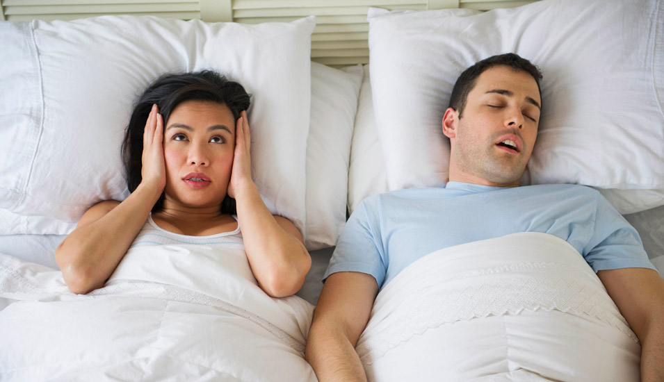 Ein Paar liegt im Bett. Da er schnarcht, hält sie sich die Ohren zu. 