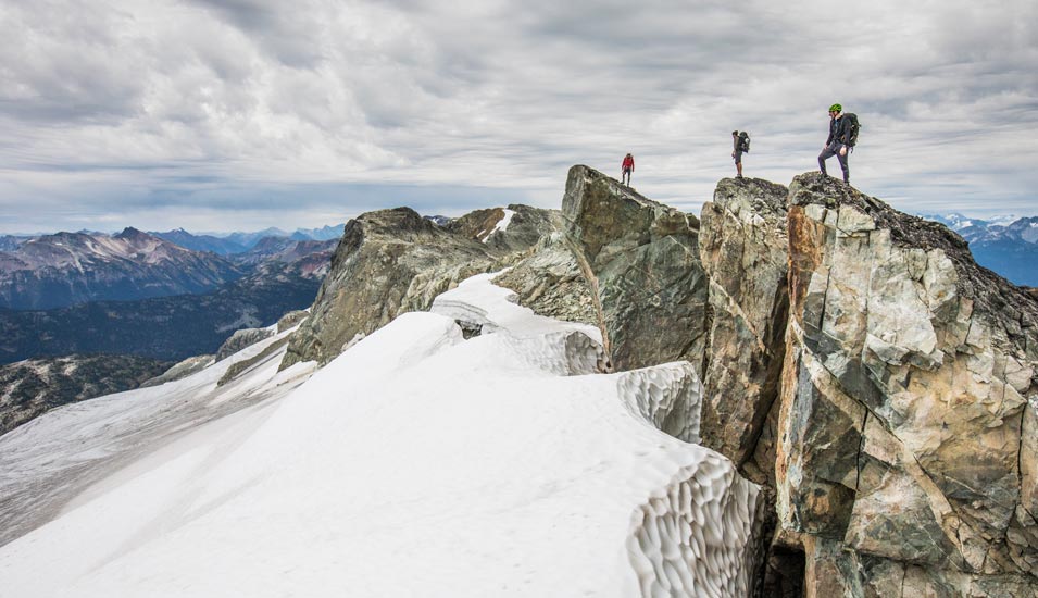 Auf drei Bergspitzen sind drei Menschen zu sehen, die in die ferne Berglandschaft blicken. 