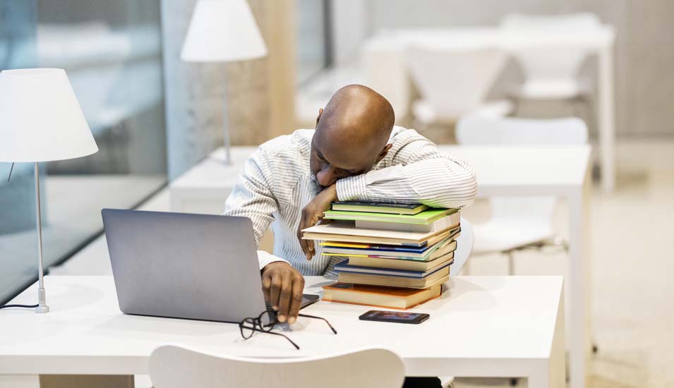 Mann mittleren Alters lehnt schlafend auf einem Stapel Bücher neben einem Laptop auf seinem Schreibtisch
