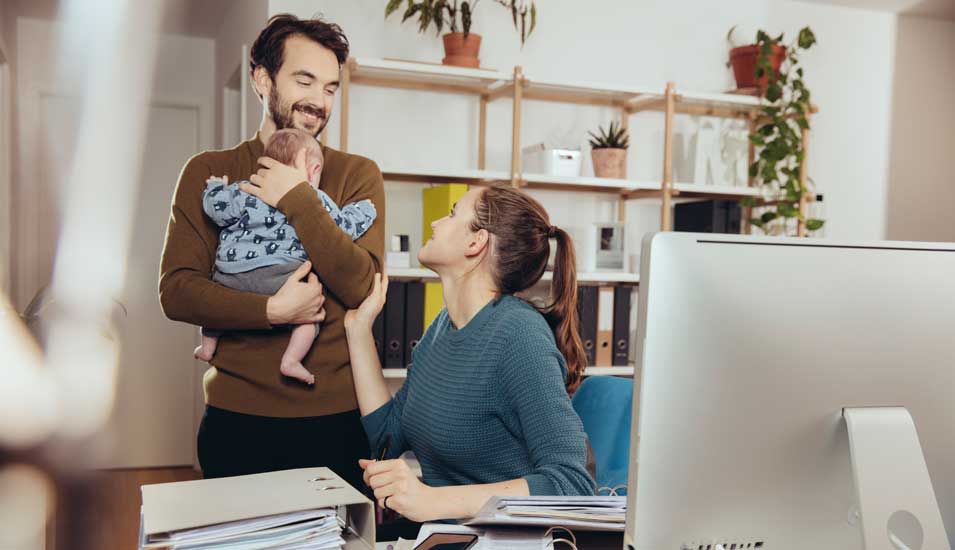 Eine Frau sitzt an einem Schreibtisch, hinter ihr steht ihr Partner und hält ihr Baby, sie lächeln sich an.