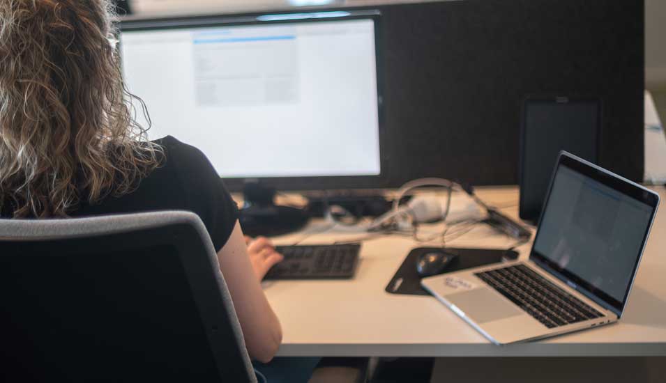 Eine Frau arbeitet gleichzeitig an einem Computer und einem Laptop, man sieht sie nur von hinten.