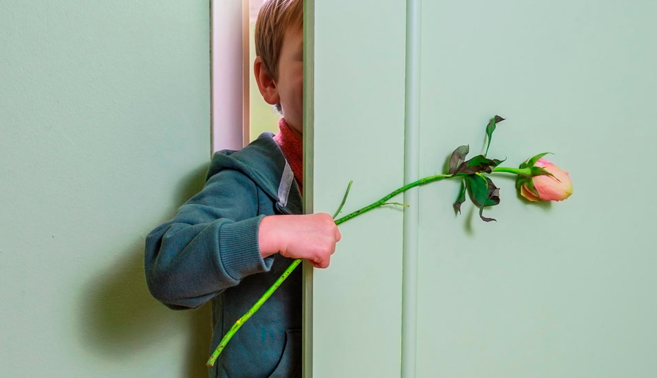 Ein Kind steht hinter einer Tür und hält eine Rose, als wolle es sich entschuldigen.