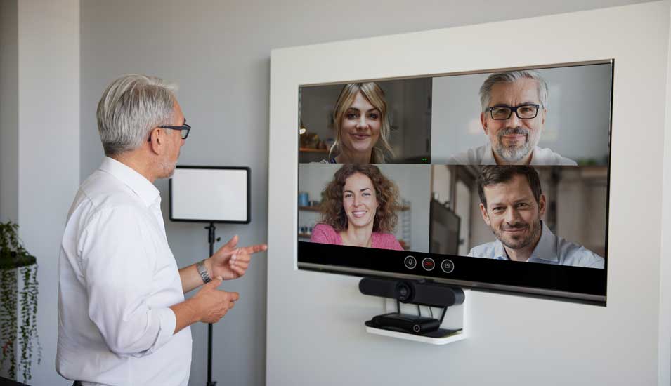 Älterer Mann unterhält sich per Videokonferenz über den Fernseher im Büro mit vier Personen, die in die Kamera blicken