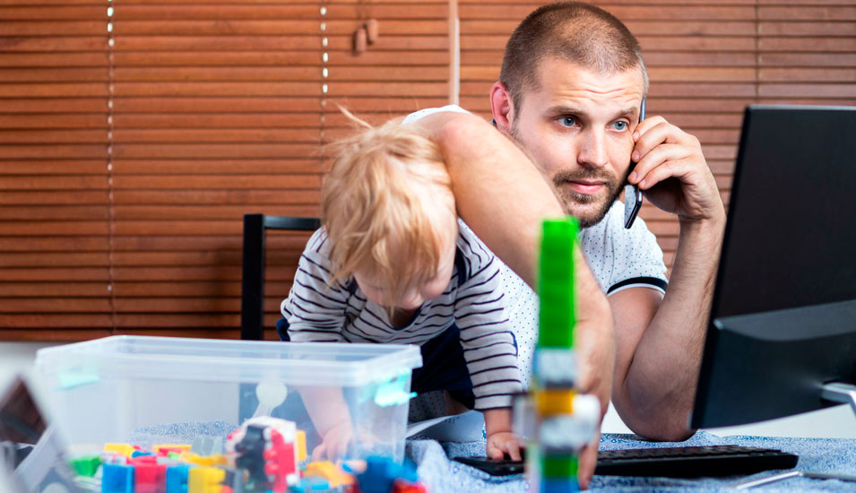 Ein Vater sitzt am Schreibtisch vor dem Computer und telefoniert, während sein Kind in seinen Armen turn und mit Legospielzeug auf dem Tisch spielt. 