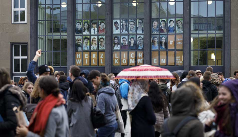 Das Foto zeigt den Haupteingang der Universität zu Köln mit großen Fotos von Studierenden und vielen Menschen.