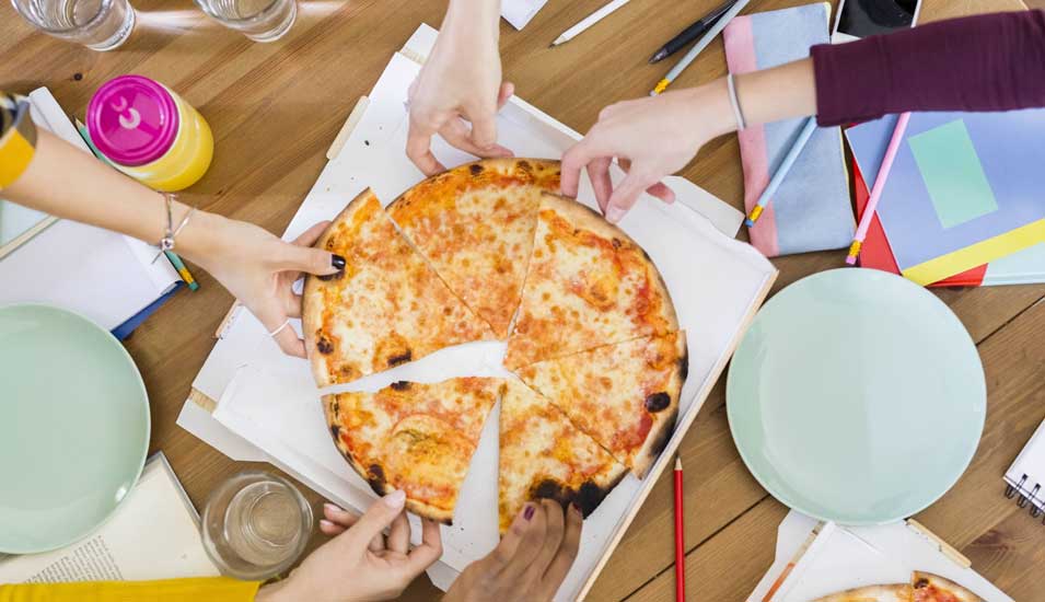 Das Foto zeigt einen Esstisch mit einer Pizza, auf die viele Hände zugreifen, von oben.