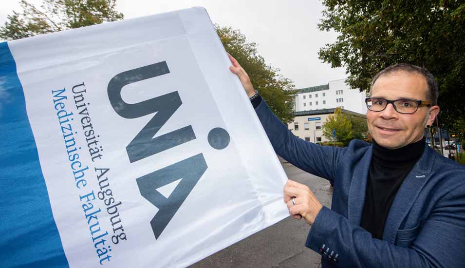 Jörn Böke, Geschäftsführer der medizinischen Fakultät der Universität Augsburg, hält vor dem Lehrgebäude ein Banner mit dem Logo und Schriftzug der Fakultät. 