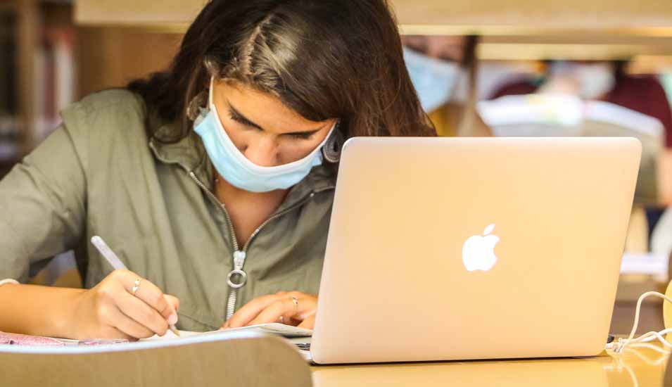 Studentin mit Mundschutz lernt während des Corona-Semesters mit Laptop in einer Bibliothek