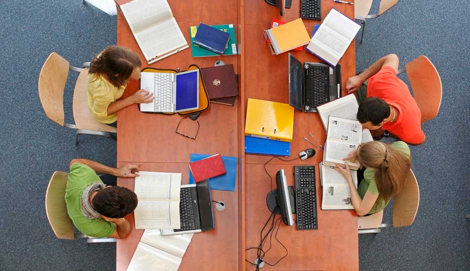 Vier Studierende arbeiten mit Laptops und Büchern in einer Bibliothek