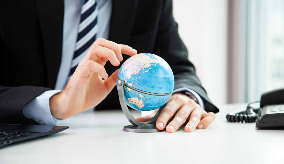 junger Mann im Anzug an einem Schreibtisch hält einen kleinen Globus in der Hand