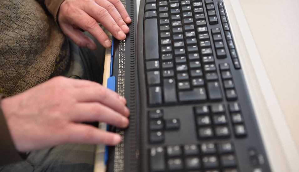 Das Bild zeigt Hände auf einer Braillezeile und eine Computertastatur.