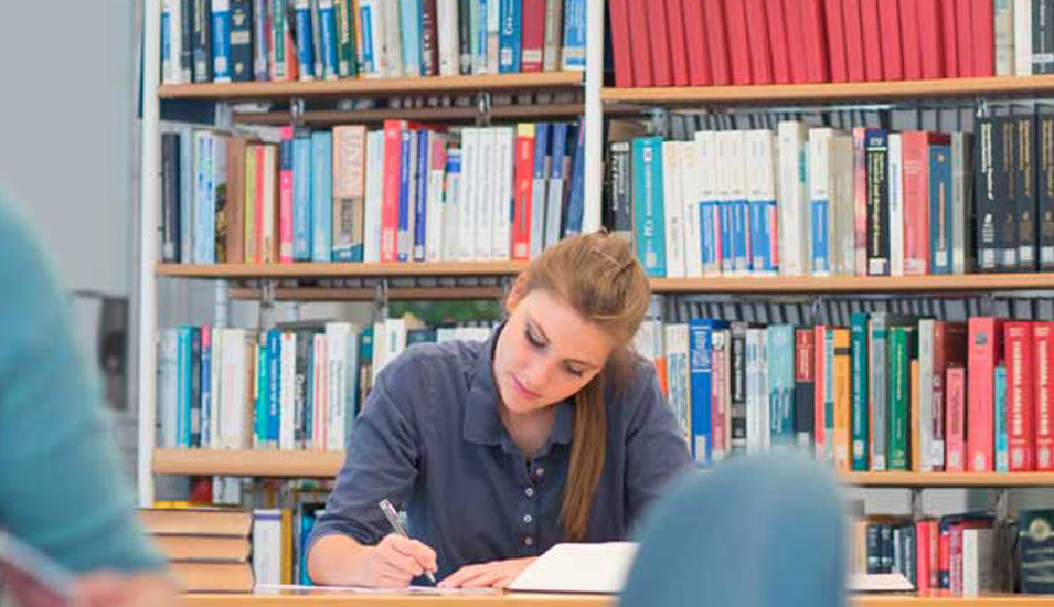 Studentin sitzt in der Bibliothek und lernt. Im Hintergrund steht ein Regal mit Lehrbüchern.