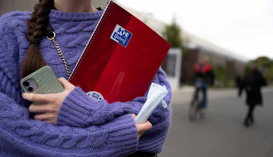 Eine Studentin steht im Strickpullover mit Handy, Block und Maske in der Hand auf dem Gelände einer Universität.
