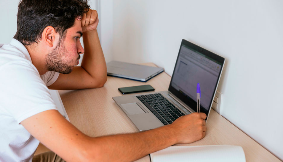 Seitenansicht eines Studenten, der vor seinem Laptop an einer Online-Klausur sitzt und angestrengt ausatmet.
