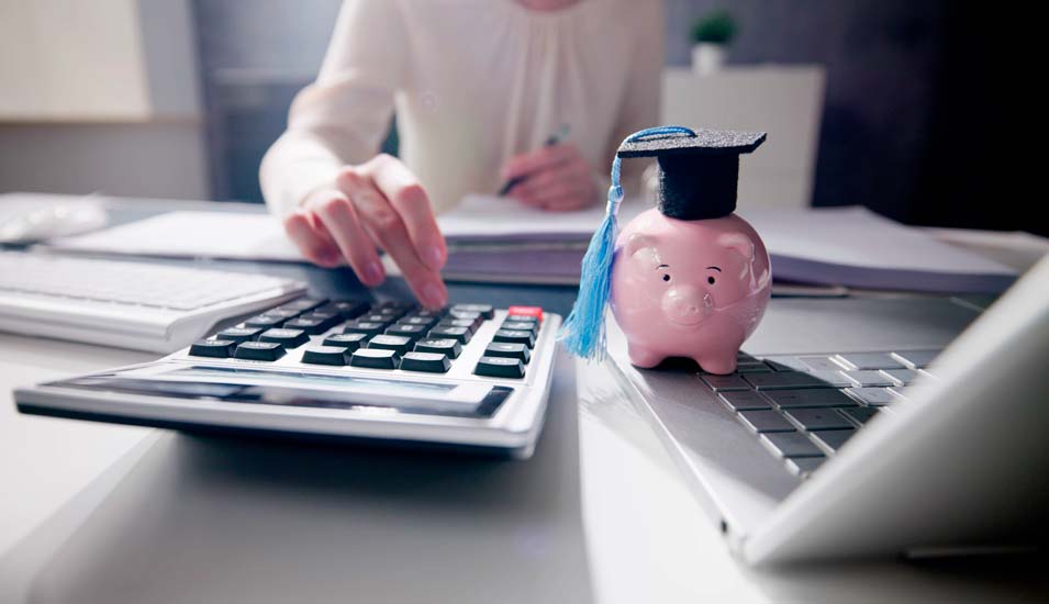 Symbolbild für einen Studienkredit: Eine junge Frau tippe auf einem Taschenrechner, daneben steht ein Sparschwein mit einem Doktorhut