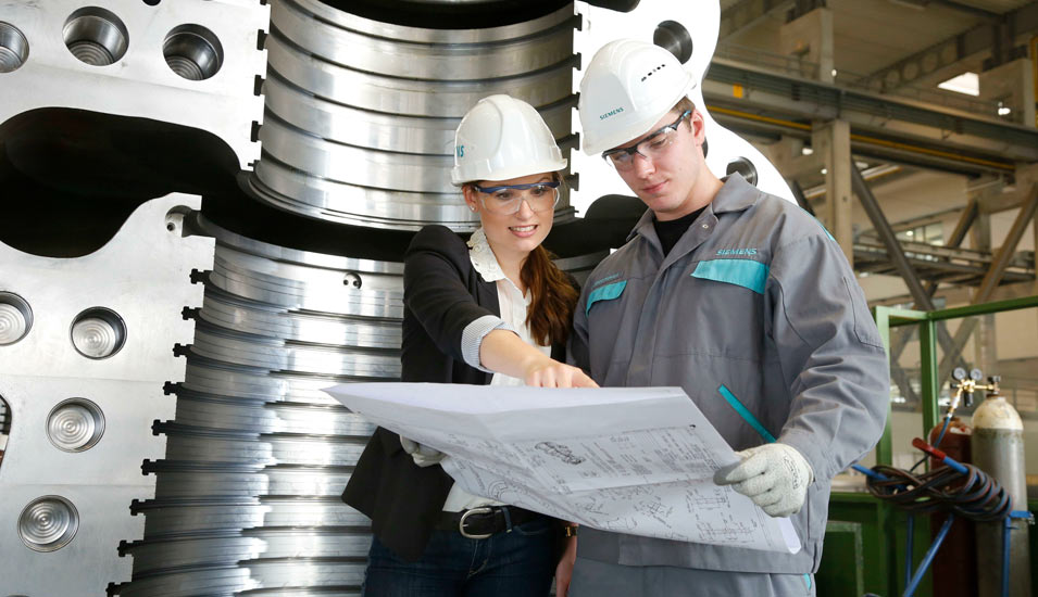 Eine junge Ingenieurin und ein junger Ingenieur stehen in einer großen Maschinenhalle und beraten gemeinsam über einen großen Konstruktionsplan. 