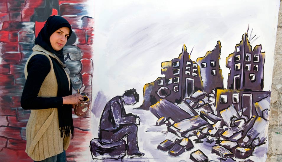 Eine junge Frau steht neben einer Häuserwand, auf die sie ein zerstörtes Gebäude gemacht hat in grau-schwarzen Farben. 