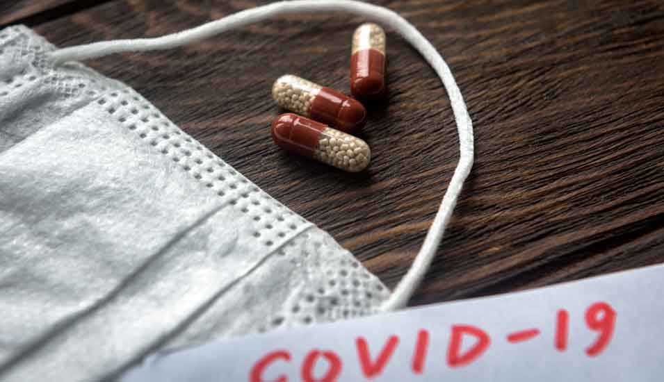 Atemschutzmaske und Medikamentenkapseln neben einem Zettel mit der Aufschrift "Covid-19"