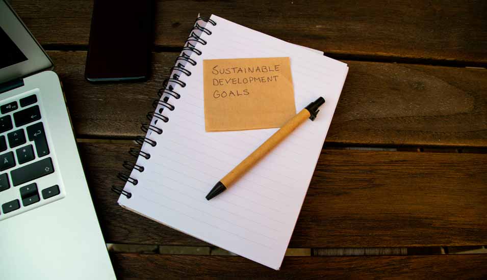 Notizblock und Stift liegen neben einem Laptop, auf dem Notizblock steht Sustainable Development Goals geschrieben