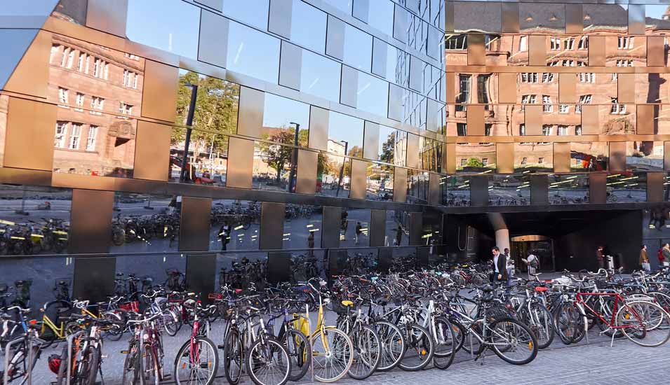 Fahrräder vor der Universitätsbibliothek Freiburg, in deren Glasfassade sich das Unigebäude spiegelt