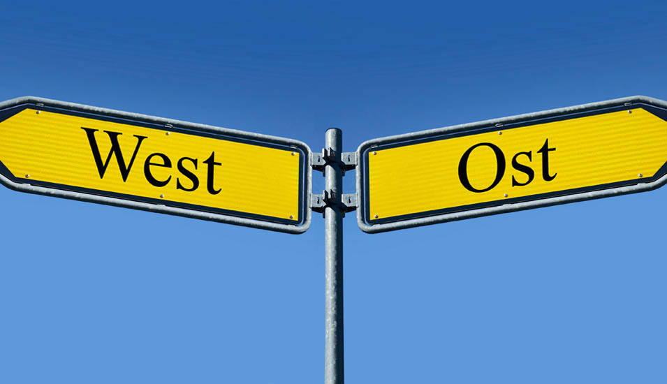 Zwei Straßenschilder zeigen in entgegengesetzte Richtungen. Auf einem steht West, auf dem anderen Ost.