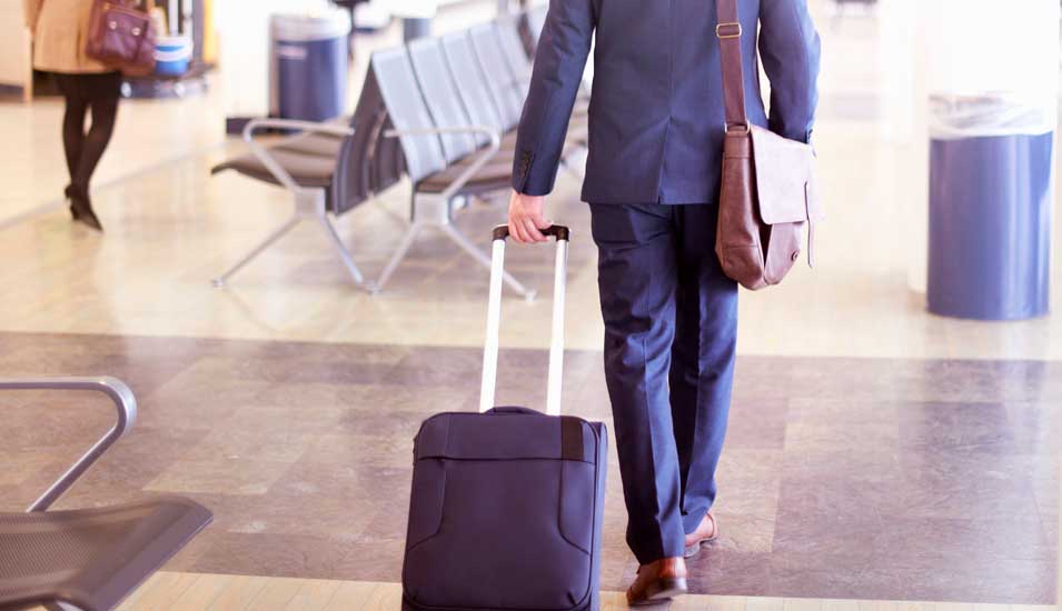 Das Foto zeigt einen Mann mit Rollkoffer auf einem Flughafen vor dem Check in.