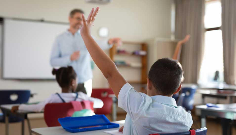 Bild eines Klassenzimmers, vorne steht ein Lehrer und ein Junge meldet sich.