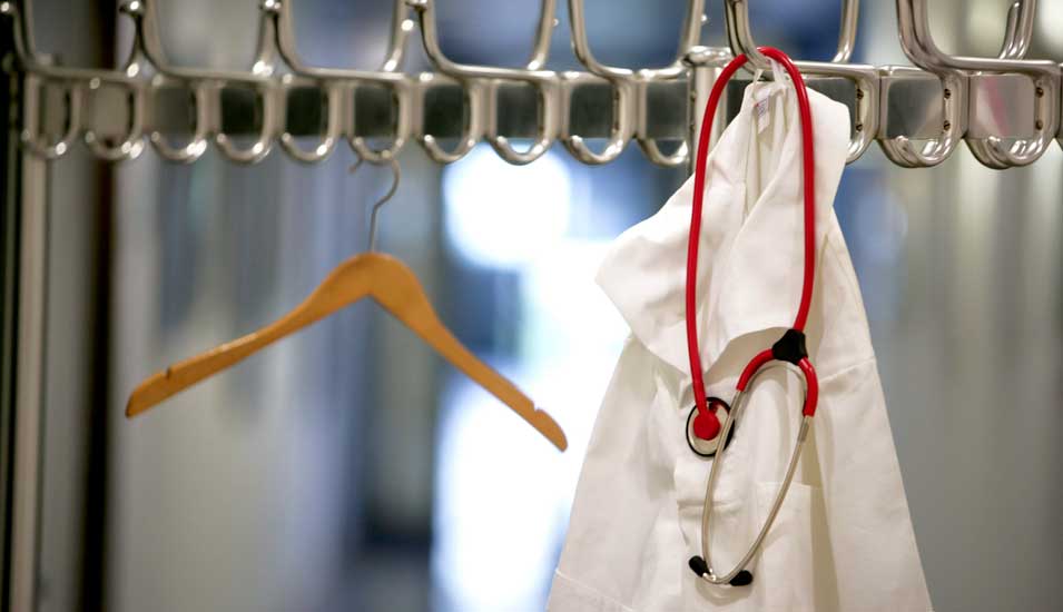 Ein weißer Arztkittel und ein rotes Stethoskop hängen an einem Garderobenständer.