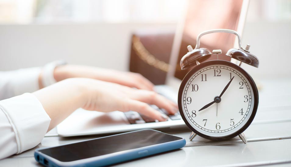 Arbeitszeiterfassung: Ein Wecker und ein Smartphone stehen auf dem Schreibtisch einer jungen Frau, die auf einem Laptop arbeitet
