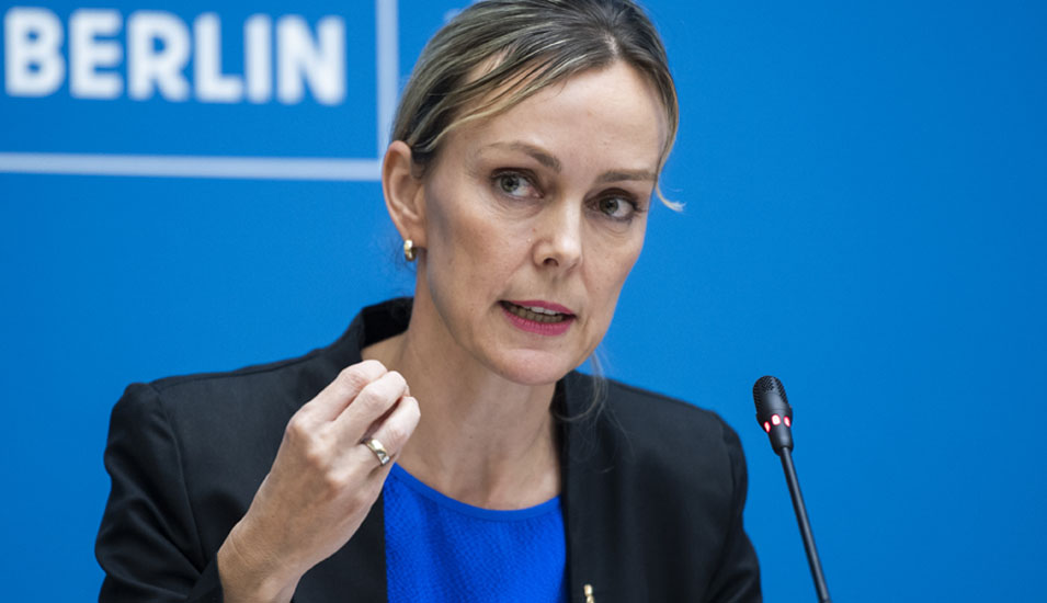 Ein Foto von Manja Schreiner, die vor einer blauen Wand stehend in eine Mikrofon spricht