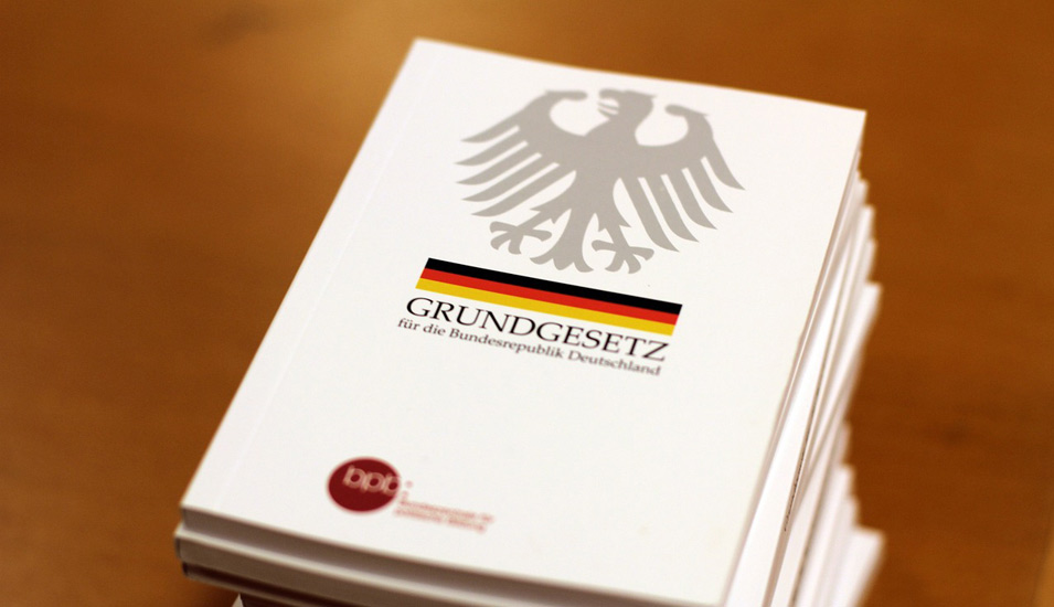 Auf einer braunen Holzfläche liegt ein Stapel von weißen, kleinen Büchern mit der Aufschrift "Grundgesetz für die Bundesrepublik Deutschland"