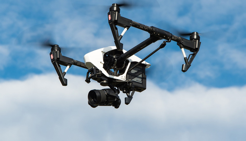 Eine Drohne mit integrierter Kamera fliegt vor einem leicht bewölkten Himmel. 
