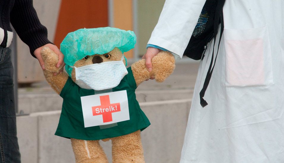Ein als Arzt verkleideter Bär mit einem Aufruck "Streik" wird von zwei Personen getragen, von denen man nur die Beine und Arme sieht. Einer davon trägt einen weißen Kittel. 