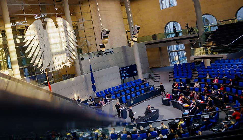 In einem großen Saal ist der Bundesadler an der Wand zu sehen und Abgeordnete sitzen auf den blauen Stühlen des Deutschen Bundestages. 