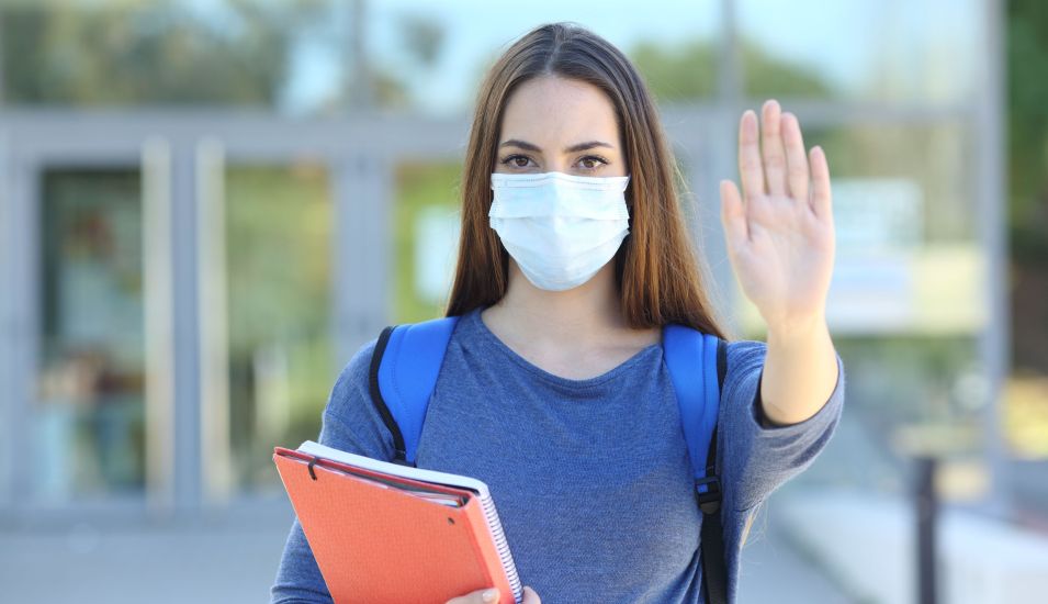 Studentin mit Unterlagen in der Hand und Atemschutzmaske gibt mit der Hand ein Stopzeichen