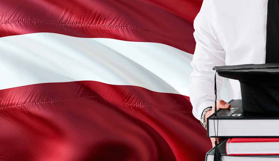 Junger Mann in Hemd und Krawatte steht vor der lettischen Flagge und hält einen Stapel Bücher und einen Doktorhut
