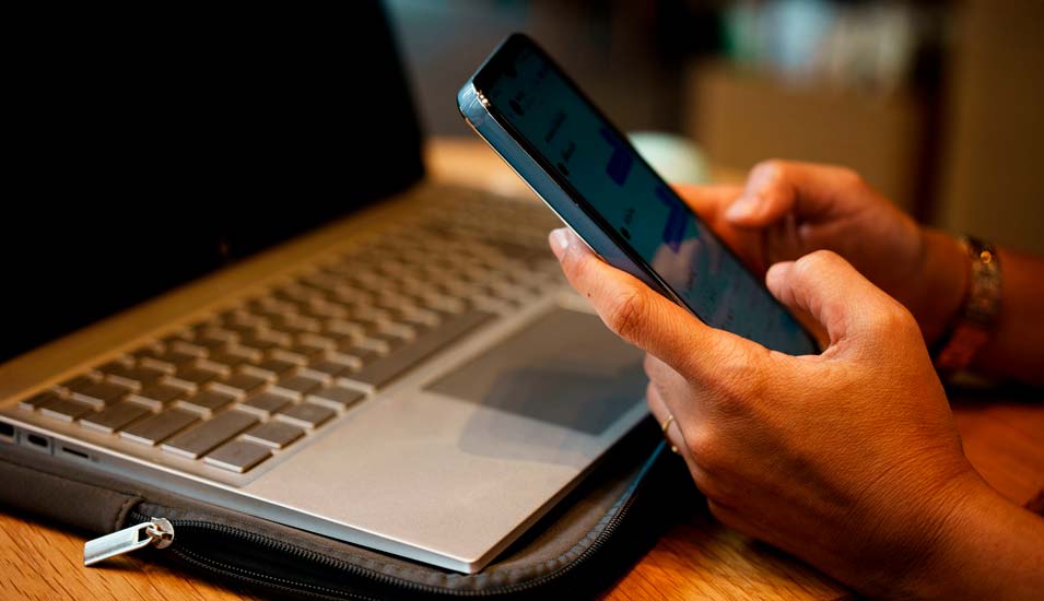 Nahaufnahme von Händen einer jungen Frau, die auf einem Smartphone chattet, während sie an einem Laptop arbeitet