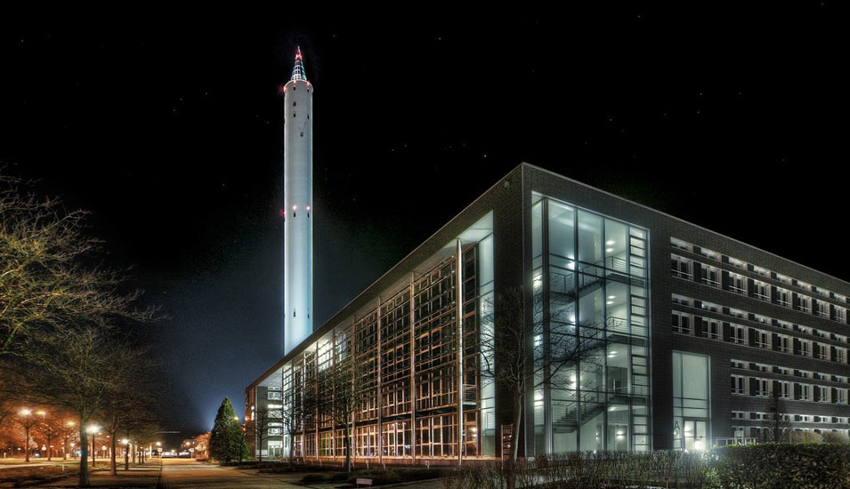 In einem Nachtmotiv ist ein großer Turm und ein großes, modernes Universitätsgebäude zu sehen. 