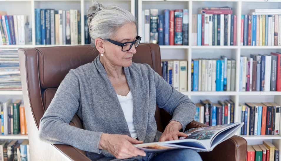 Ältere Frau sitzt in einem Sessel vor Bücherregalen und liest in einem illustrierten Buch