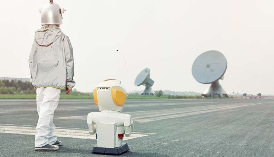 Als Roboter verkleideter Mensch und ein Roboter stehen auf einem Rollfeld mit Satellitenschüsseln