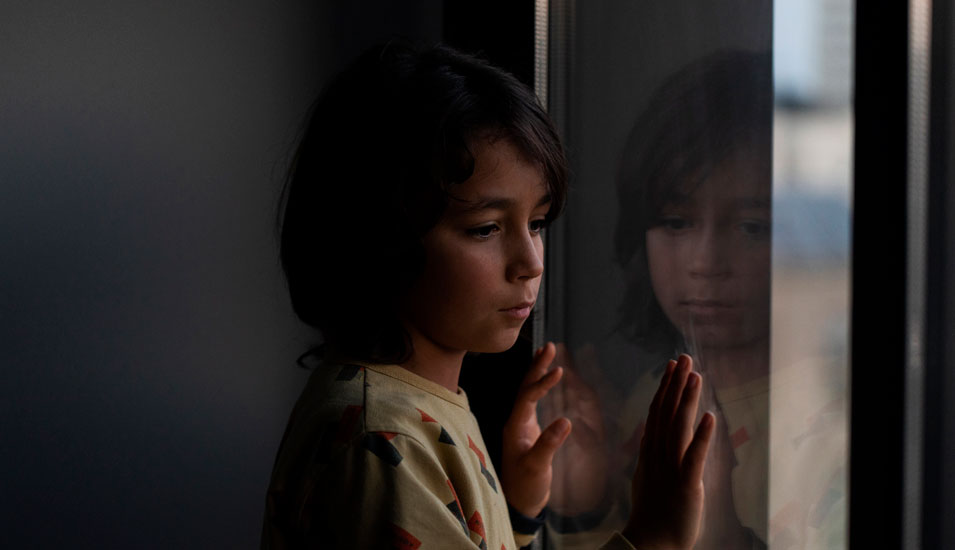 Ein Kind steht in einem dunklen Raum am Fenster und schaut nach draußen.