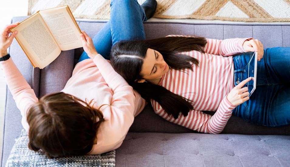 Zwei Frauen lesen auf dem Sofa, eine ein Buch, eine am Tablet
