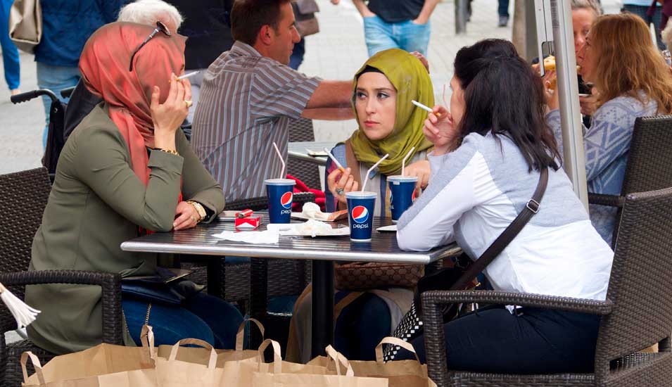 Muslimische Frauen sitzen in Deutschland an einem Cafétisch, trinken Pepsi und rauchen. Zwei von ihnen tragen ein Kopftuch.