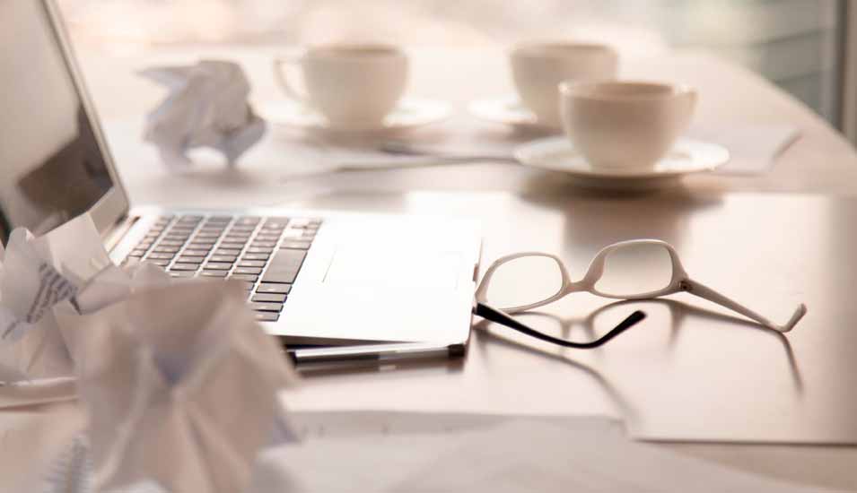 Arbeitsplatz mit Laptop, mehreren Tassen Kaffee, einer Brille und zerknülltem Papier