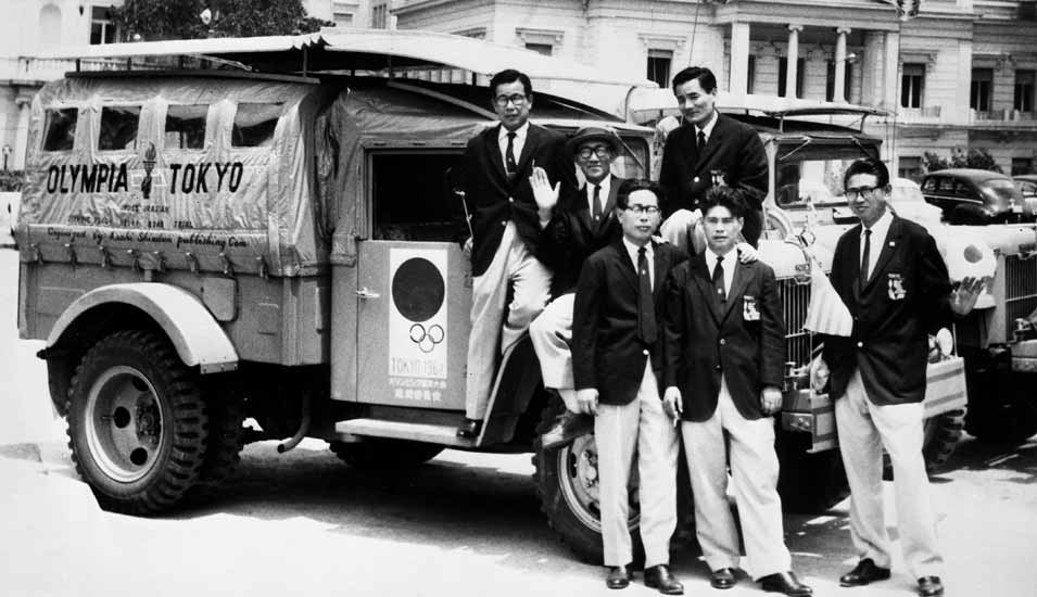 1964: Gruppe von japanischen Athleten vor einem LKW mit Logo der Olympischen Spiele beim Abholen des Feuers in Athen für die Spiele in Tokio.