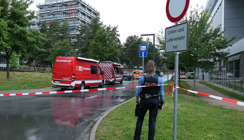 Blick auf das abgesperrte Gelände rund um Gebäude L201 der TU Darmstadt mit Einsatzwagen der Feuerwehr und einer Polizistin.