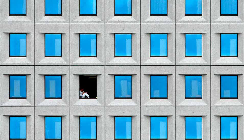 Gebäudefassade mit zahlreichen identischen geschlossenen Fenstern und einer Person, die in einem geöffneten Fenster sitzt