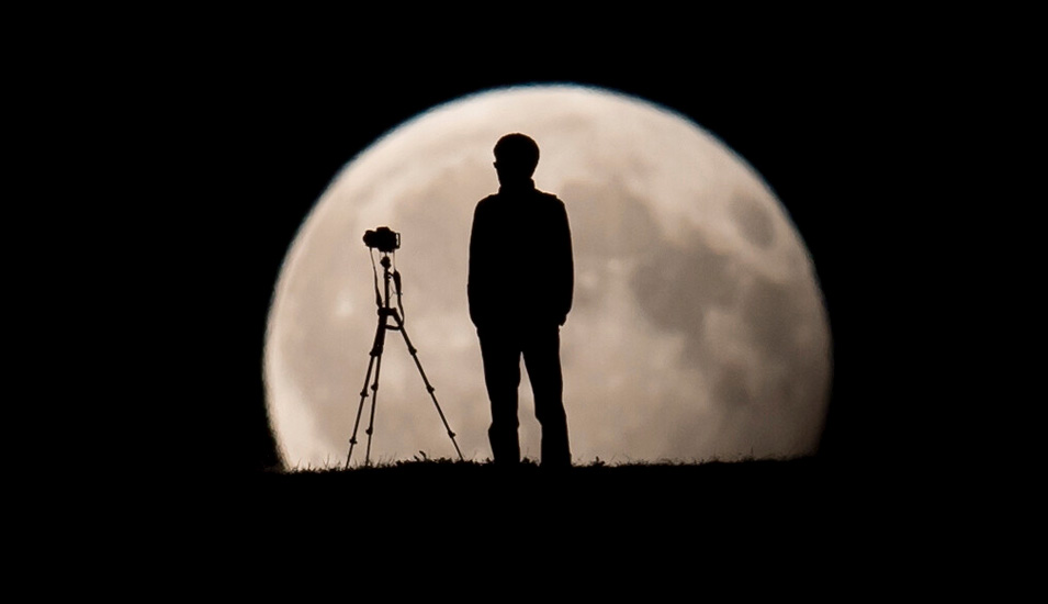Ein Mann fotografiert am den Mond, er ist als dunkle Silhouette vor dem hellen Mond zu sehen.