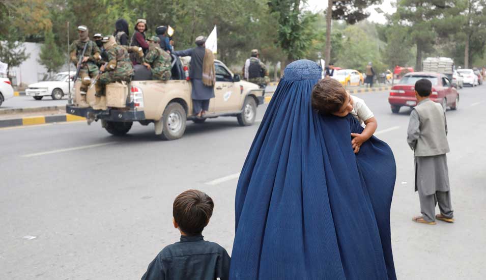 Straßenansicht in Kabul vom 15. August 2022: Im Bildvordergrund sieht man eine Frau von hinten mit einem Kind auf dem Arm, im Bildhintergrund fährt ein Pick-Up-Truck voller Taliban-Kämpfer vorbei.