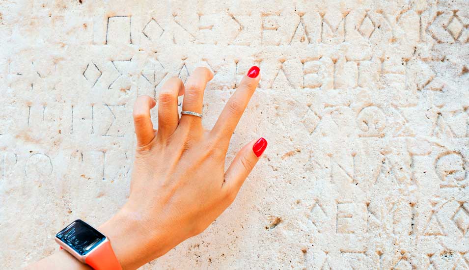 Hand einer jungen Frau, die auf die Schrift in einer antiken, griechischen Ruine zeigt und versucht die Schrift zu entziffern.